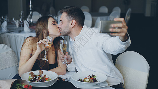 社交在餐厅吃晚餐时 迷人的情侣正在使用智能手机与香槟酒杯自拍 他们微笑着 亲吻着 摆着姿势看着镜头背景