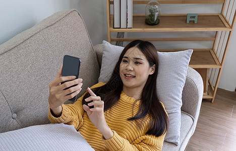年轻的亚洲女性正在沙发上使用智能手机 放松 快乐 技术概念女孩自由闲暇电话房间微笑耳机说谎音乐房子图片