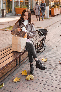 浓情咖啡一个时尚的黑发女人在秋天的城市里走来走去 黑发女人坐在外面的长凳上 手里拿着一杯咖啡和一部电话 手里拿着一个带法式面包的工艺包情背景
