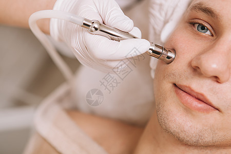 在美容院接受皮肤护理治疗的英俊年轻男子青年程序服务仪器去角质剥皮医生皮肤科诊所男人背景图片