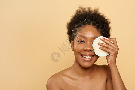美国黑人年轻女性在她的眼睛附近拿着一个可重复使用的护肤棉垫 微笑着看着镜头 零废物概念图片