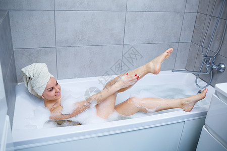 头戴白毛巾的女孩躺在白色浴缸里的浴室里 手上沾了很多浑浊的肥皂 全漩涡泡沫 辛苦一天后放松一下 水疗放松治疗身体乐趣温泉气泡说谎图片