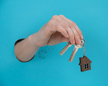 女人的手握着钥匙 钥匙链以房子的形状 在蓝色背景上饰品销售代理人钥匙圈保险公寓贷款女性抵押金融图片