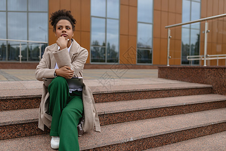 在商业大楼的背景下 一位年轻的非洲裔美国女性穿着职业装坐在台阶上 若有所思地凝视着远方图片
