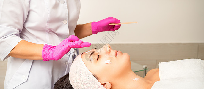戴着手套的美容师在女性脸上涂上带去角质霜的保湿面膜 面部美容治疗 面部护理程序面具摄影皮肤清洁诊所卫生沙龙老化滋润按摩图片