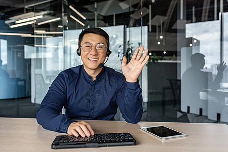 网络摄像头视图 成功的技术支持和客户服务人员的肖像 男人微笑着看着镜头挥手打招呼 在现代办公楼内工作的亚洲人图片