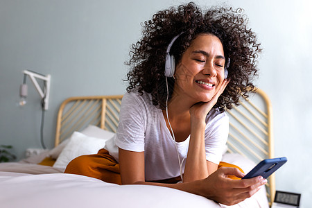 年轻多种族妇女利用耳机和手机在家中舒适卧室的床上享受音乐 并在床上放松图片