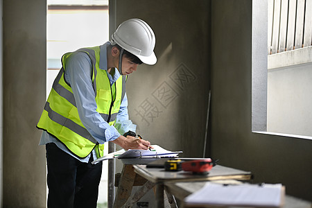 身着安全头盔和防弹背心的工程师男子与建筑施工工地蓝图一起工作图片