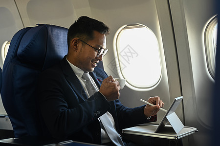 飞机撒药微笑的商务人士喝咖啡 使用平板药和在商务旅行飞行期间使用平板电脑背景