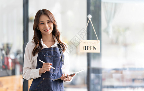 淘宝店铺招牌美丽的亚洲年轻咖啡师女人穿着围裙拿着平板电脑 站在咖啡馆门前 上面有开放的招牌 企业主创业理念咖啡营销员工拉丁工人食堂热饮药片职背景