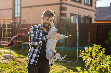 年轻男子在后院的家附近拿着杰克鲁塞尔特瑞狗     宠物主和动物友谊猎犬场景闲暇行动斑点植物喜悦微笑衬衫图片
