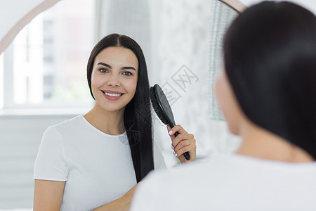 一位美丽的年轻女士站在镜子里看着自己 用梳子梳头发 她把头发梳了一下衬衫女性发型刷子浴室成人派对护理黑发微笑图片