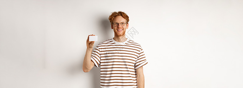 界面展示购物和财务概念 微笑的年轻人 长着鬃毛和红头发 展示塑料信用卡 看起来很开心 白色背景信用促销发型广告冒充银行胡子眼镜红色卡片背景