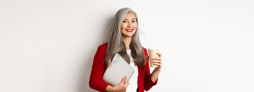 商业 头发灰白 穿着红色西装外套 喝咖啡 手拿笔记本电脑站立 白色背景的成功亚洲女商务人士咖啡套装经理电子商务化妆品企业家办公室背景图片
