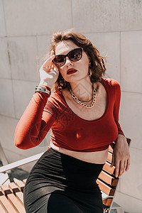 一个女人在街上的画像 外面的长凳上坐着一个戴着眼镜 穿着红色衬衫和黑色裙子的迷人女人姿势城市人行道微笑冒充条纹女孩连衣裙购物头发图片