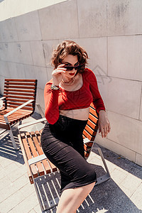 一个女人在街上的画像 外面的长凳上坐着一个戴着眼镜 穿着红色衬衫和黑色裙子的迷人女人腰带卷曲女孩头发冒充购物成人微笑条纹女性图片