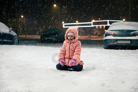 快乐的孩子 穿着暖冬工装裤的小女孩坐在雪街上 在晚上散步时享受第一场雪图片