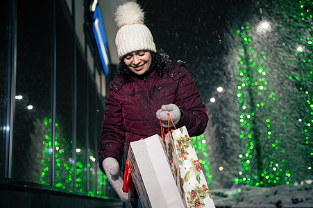 迷人的女人拿着购物袋 走在街上 在冬天的雪之夜被节日照明照亮了图片