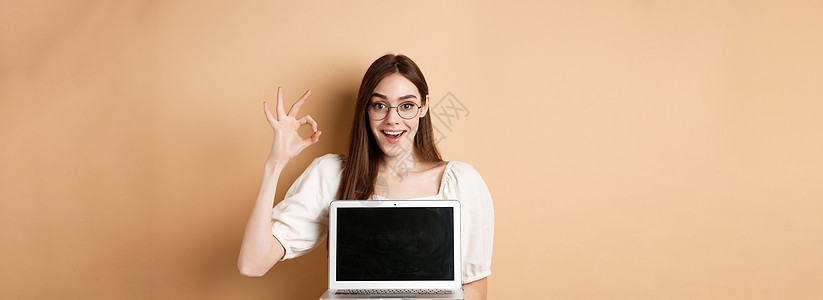 身戴眼镜 显示好手牌和笔记本电脑屏幕 建议互联网宣传 站在蜜蜂背景上兴奋的年轻女青年女性技术广告褐色学生眼镜微笑纪念日女孩工作图片