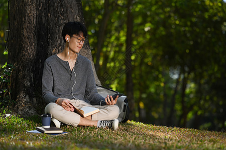 在阳光明媚的美日 在绿草上用树下听耳机和移动电话监听音乐并使用绿色青草图片