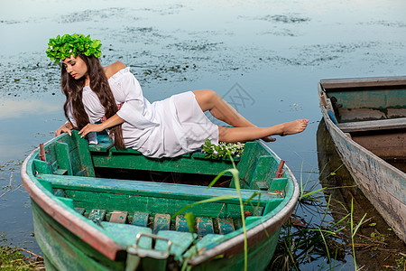 深色头发长发的女神 穿着白色古董礼服 坐在河中央的一条船上神话裙子冒充童话美人鱼魔法魅力若虫花朵女孩图片