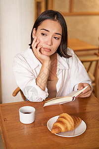 对亚洲女孩的垂直拍摄 坐在咖啡馆里独自一人 阅读书本 看起来很沮丧 喝着咖啡和羊角面包企业家女性午餐酒吧学生作家电脑早餐商务上网图片