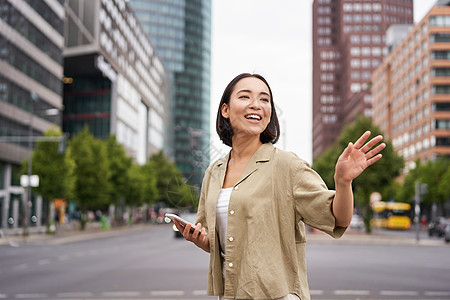 快乐的亚洲女孩路过朋友 在街上挥手向她们挥手 在城市散步时打招呼 拿着智能手机女性青少年电话街道技术学生黑发细胞互联网出租车图片