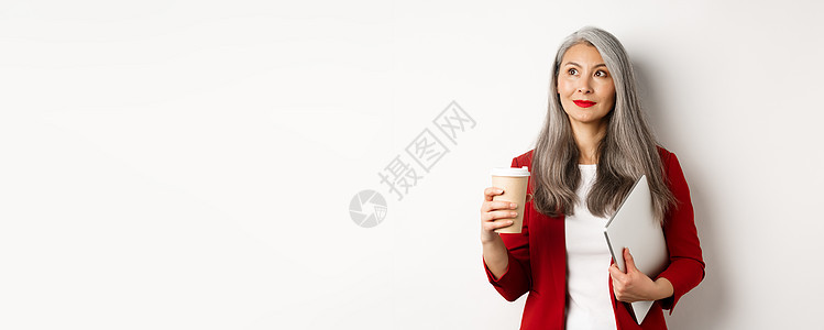 成功的亚洲商业女商务人士 灰发 喝咖啡和用笔记本电脑站立 看深思熟虑的左上角工作电子商务化妆品成人西装人士咖啡祖母自由职业者头发背景图片