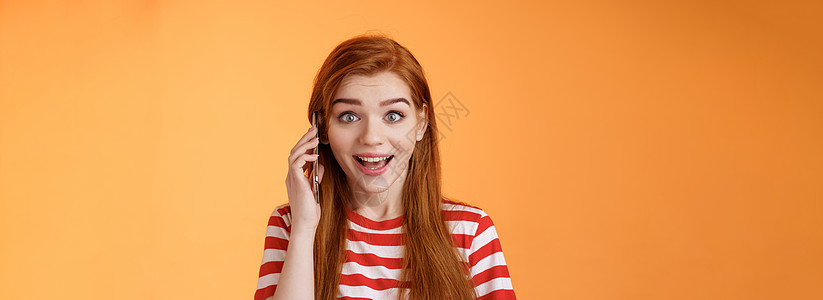 快乐听到朋友的声音 在国外打电话 逗乐的可爱的红发快乐女孩拿起电话 拿着智能手机的耳朵 发现好消息 微笑着惊讶 站着橙色背景兴奋图片