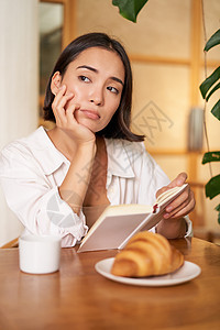 对亚洲女孩的垂直拍摄 坐在咖啡馆里独自一人 阅读书本 看起来很沮丧 喝着咖啡和羊角面包上网学生人士企业家商务黑发女性潮人午餐技术图片