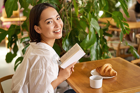 美丽的亚洲年轻女性手里拿着一本书 坐在咖啡馆里 喝着咖啡 吃着羊角面包 微笑着 看起来很神秘上网情感午餐网络早餐笔记本商务商业作图片