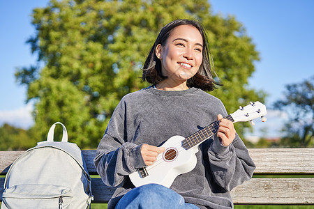 可爱的年轻女子坐在阳光明媚的公园长椅上 背着背包坐在书架上 弹四轮吉他和歌唱喜悦音乐幸福爱好音乐家快乐唱歌乐器女性微笑图片
