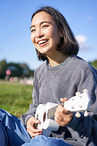 微笑的亚洲女孩唱歌和笑 弹四弦琴 学习弹奏乐器 坐在公园户外的垂直镜头花园歌曲音乐家音乐黑发乐趣幸福爱好女性快乐图片