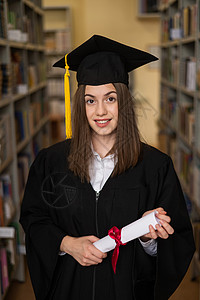 毕业学士穿着毕业礼服的快乐年轻女子 在图书馆持有文凭地幔书架教育证书成就女性庆典学校研究生法律背景