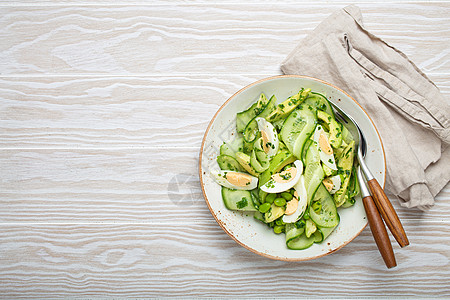 健康的绿鳄梨沙拉碗 配有煮鸡蛋 切片黄瓜 豆子 橄榄油和陶瓷板顶端的草药 白木制餐桌背景图片