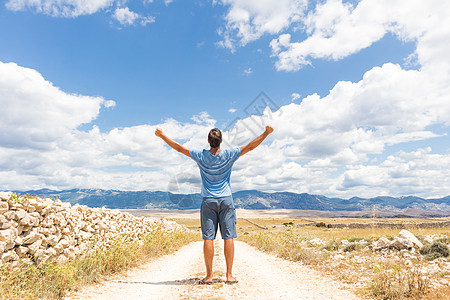 站在泥泞的乡间小路上的休闲运动男子的后视图在蓝色的夏日天空中举起手到云层 自由和旅行冒险的概念 闲暇 短裤图片