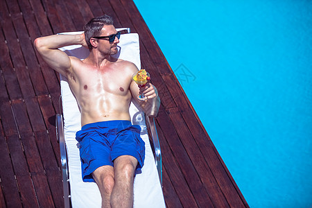男人拿着马提尼杯子 在一个遮太阳的休息者身上放松时 游泳池图片