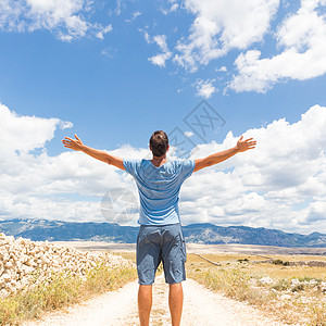 站在泥泞的乡间小路上的休闲运动男子的后视图在蓝色的夏日天空中举起手到云层 自由和旅行冒险的概念 精神 享受图片