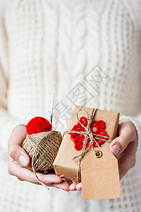 穿着白色针织毛衣和手套的女人拿着一份礼物和一卷卷红线和亚麻绳 礼品采用手工钩编红色雪花牛皮纸包装 Present 的文本有一个空图片