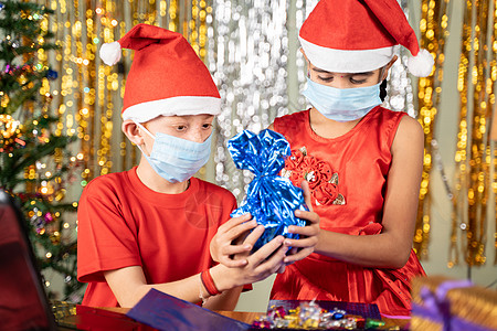 戴着医用面具的孩子们在笔记本电脑前在圣诞前夕在家里打开带有装饰背景的礼物 — 由于冠状病毒或 covid-19 大流行而导致遥远图片