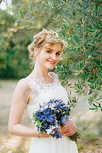 一位手拿着一束蓝花的新娘 在果林和微笑中站在绿橄榄树枝旁图片