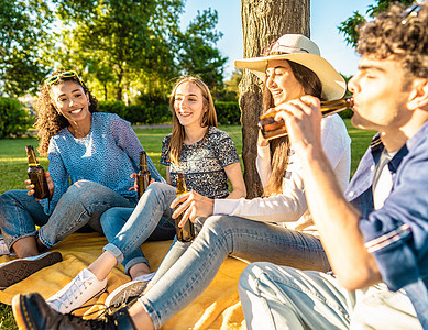快乐的朋友团体在城市公园户外喝着瓶装啤酒庆祝坐在草地上 无忧无虑的微笑的年轻人在日落时喝着酒在大自然中玩乐 在草地上野餐图片