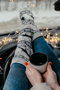 穿着袜子和袜子的女子腿手握着热水杯咖啡或冬车茶 圣诞袜 加兰灯光不亮 舒适 窗户图片