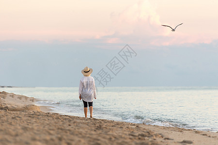 日落时在海滩上行走的妇女持有手机 戴草帽和做梦图片