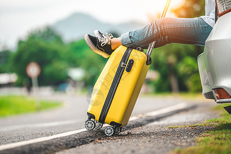 黄色行李特写镜头和放松在有路背景的汽车后面的妇女腿 公路旅行和假期假期的概念 人们的生活方式和交通 女孩在公路旅行中度过周末图片
