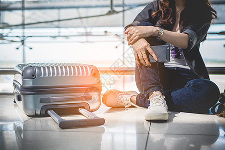 年轻女子坐在机场休息室等待出发时带着包和手提箱行李的特写 女性旅行者和旅游主题 旺季和假期的概念 放松和生活方式 互联网 门图片