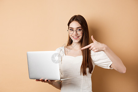 戴眼镜的微笑女性用手指指着笔记本电脑屏幕 在米色背景上展示在线宣传 假期 漂亮的图片