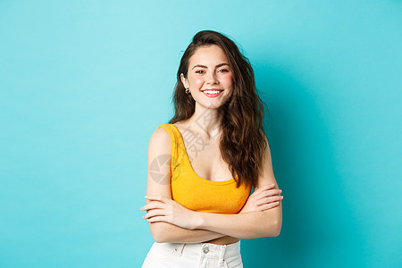 夏季和生活方式的概念 身穿黄色裁剪上衣的年轻美女 双臂交叉在胸前 对着镜头开心地微笑 站在蓝色背景下图片