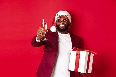 圣诞节 非洲帅美男子在聚会眼镜和圣塔帽上 持有新年礼物和香槟杯 祝愿节日快乐 红底的红色背景 胡须图片