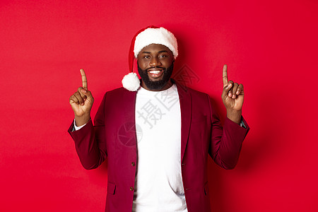 圣诞 派对和节日概念 戴着圣塔帽的非洲帅美男子微笑 举起手指并展示广告 站在红色背景上站立 笑声 促销 圣诞节图片